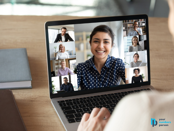 computer screen with one large woman's face and 8 smaller faces surrounding her on a zoom call, with a white woman's hand typing on the computer in the foreground bookkeeping onboarding